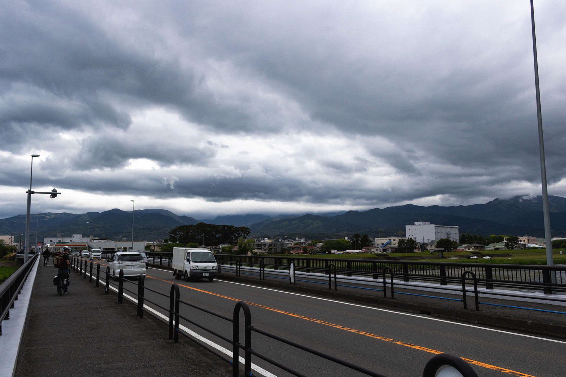 Photograph of an overcast road from the sidewalk with a cyclist futher ahead on the sidewalk.